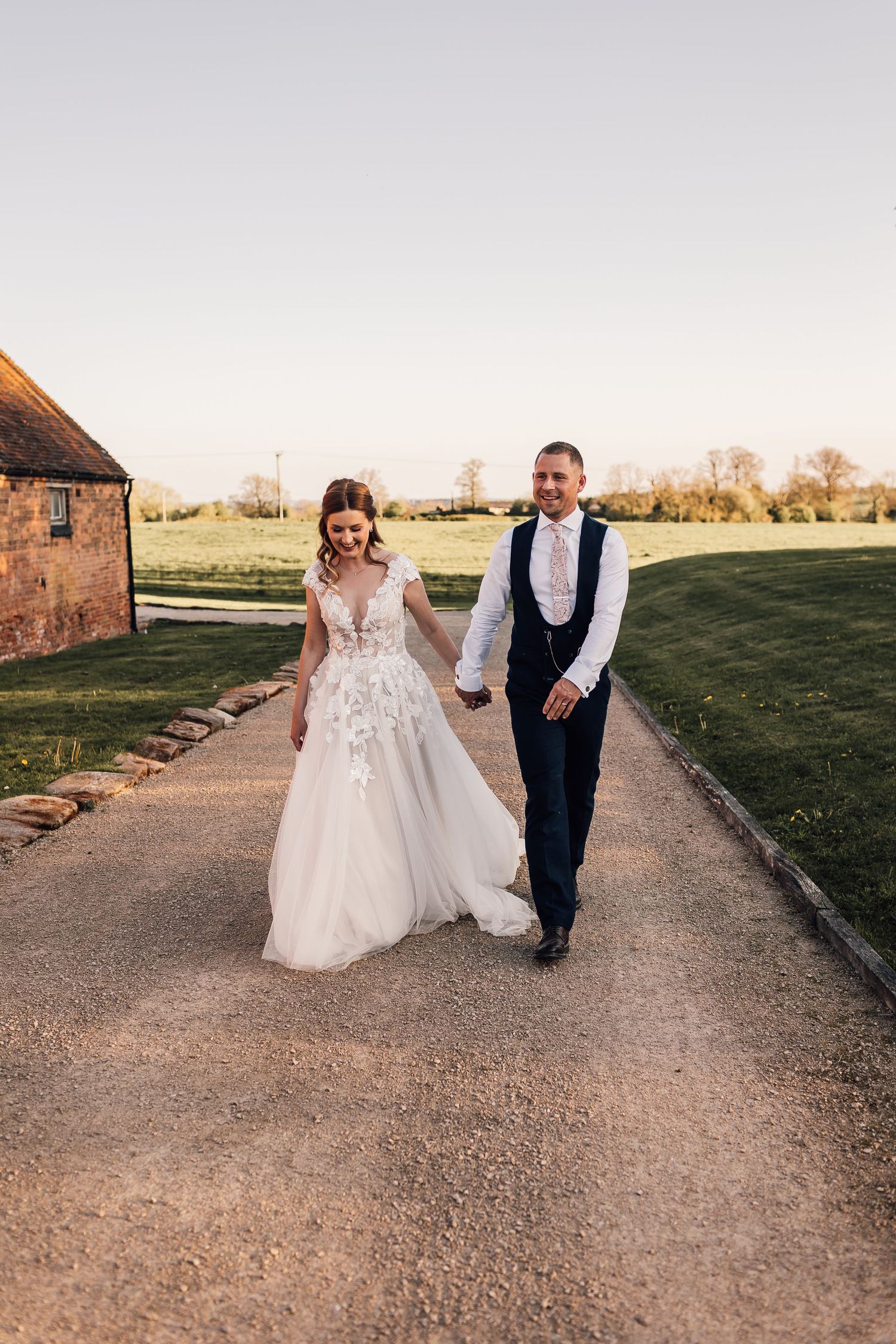 Bride and groom walk hand in hand along gravel path at golden hour outside rustic farm wedding venue.