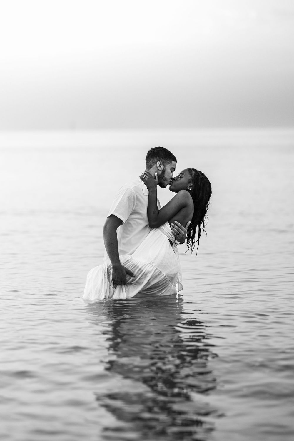A romantic couple embraces while standing in shallow ocean water in a black and white photograph.