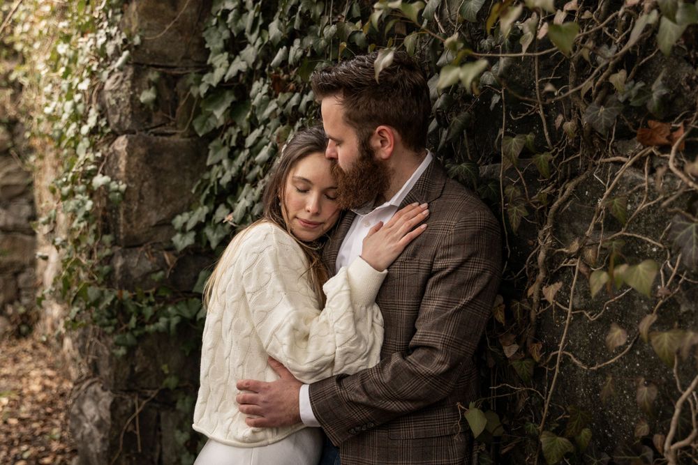 A romantic couple embraces outdoors against an ivy covered stone wall in vintage-inspired clothing.