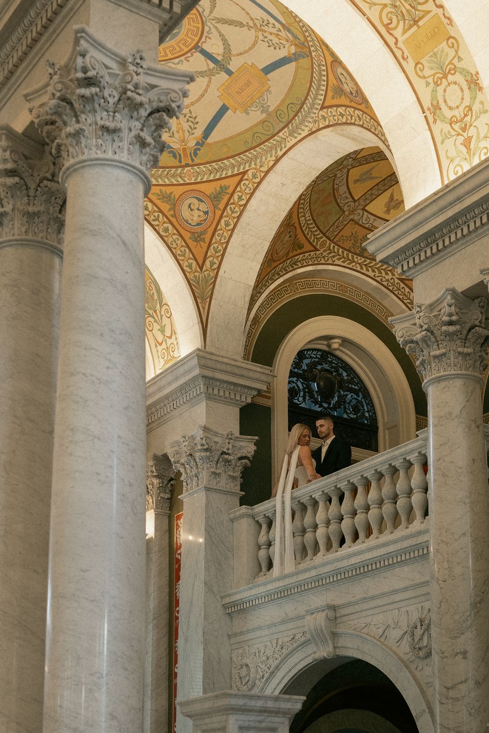 Bride and groom stand on elegant marble balcony with ornate columns and arched ceiling details.
