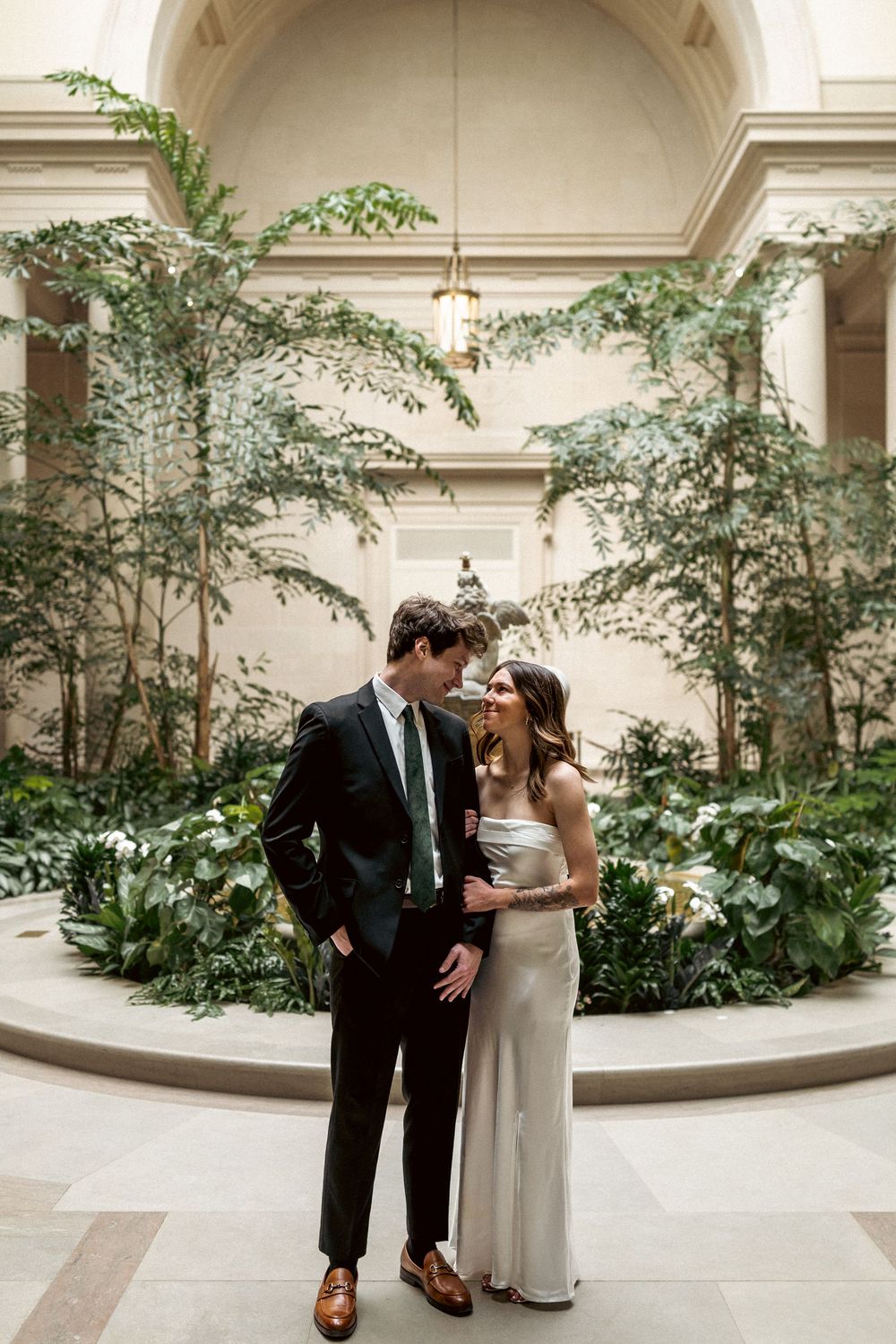 A couple shares an intimate moment in an elegant indoor courtyard with lush greenery and arched architecture.