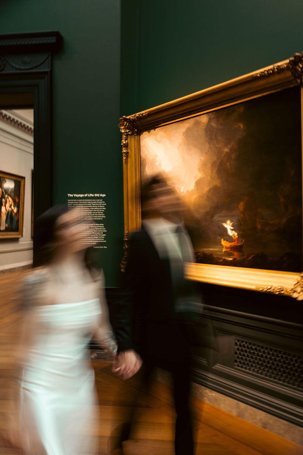Motion-blurred figures move through a dimly lit museum gallery with dark green walls and illuminated artwork displays.