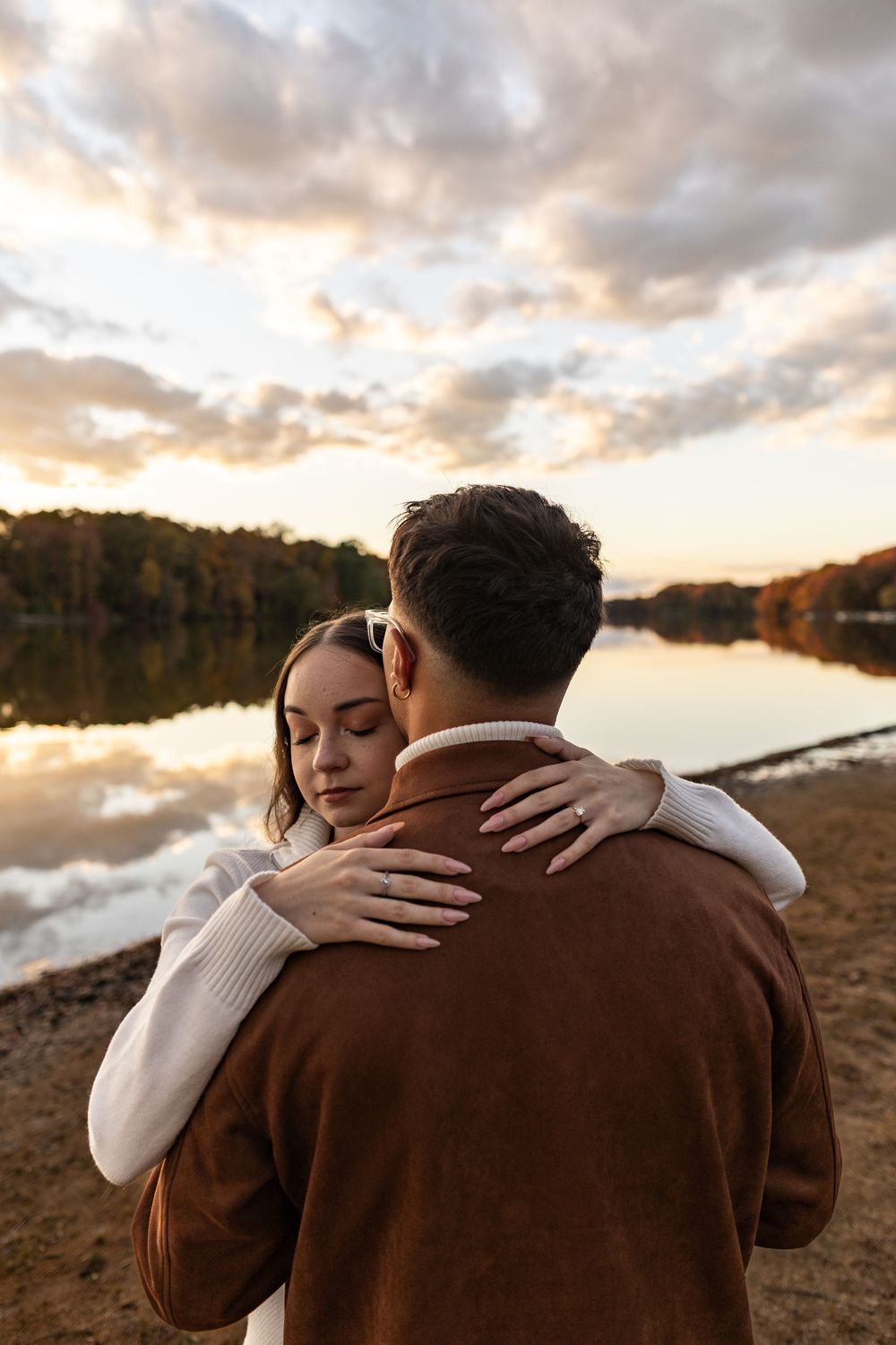 A romantic couple embraces by a serene lake at sunset with autumn trees reflecting on the calm water.