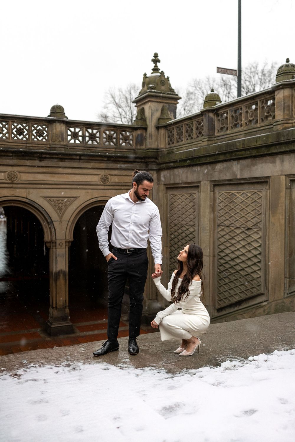 Couple in formal attire posing on snowy steps near an ornate stone bridge or terrace.