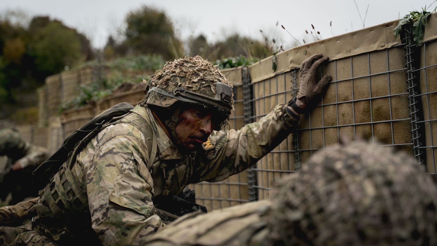 Soldier in tactical gear aims rifle amidst smoke on rocky terrain.