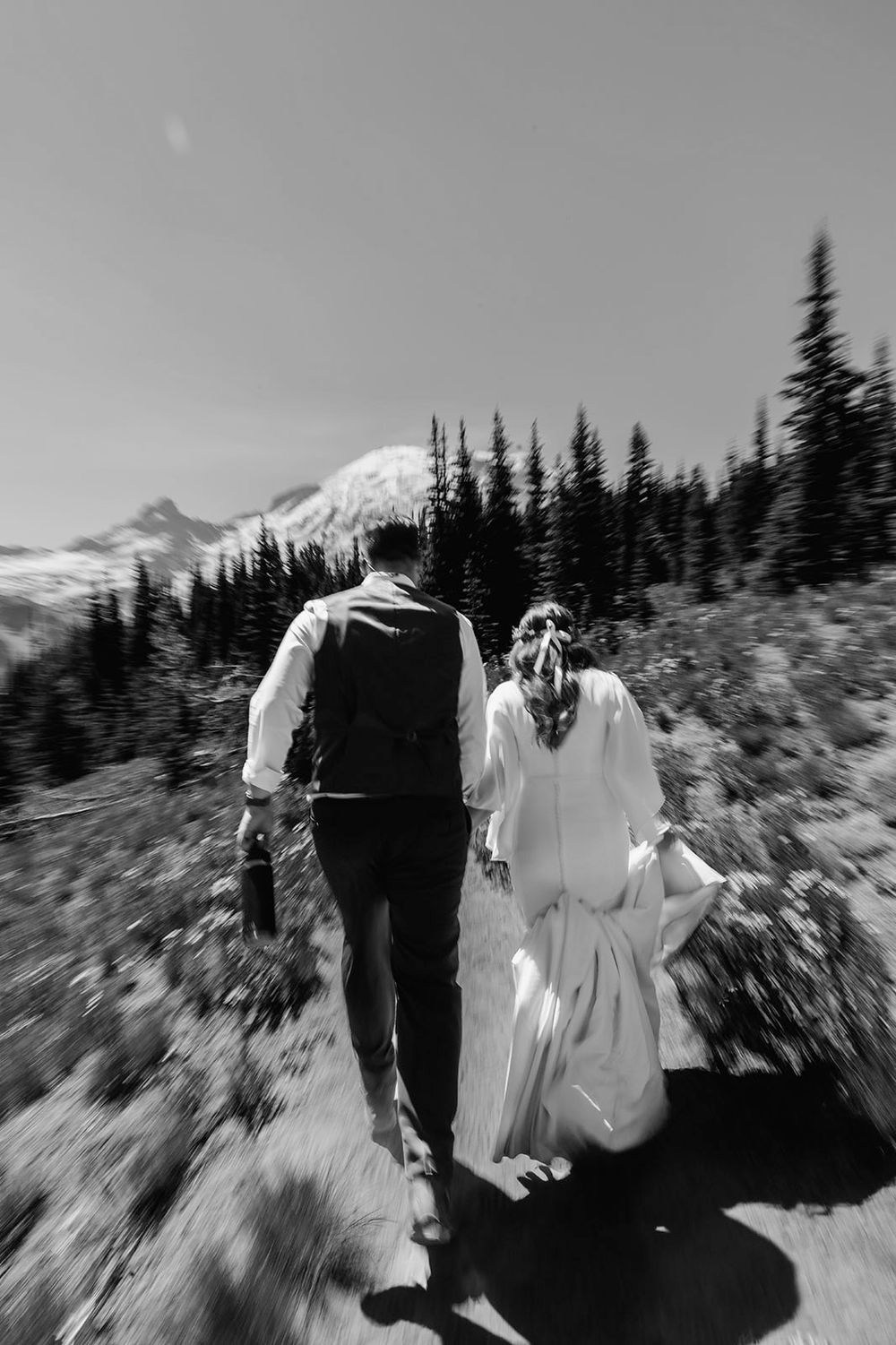 Black and white photo of bride and groom walking hand in hand toward mountain landscape with evergreen trees.