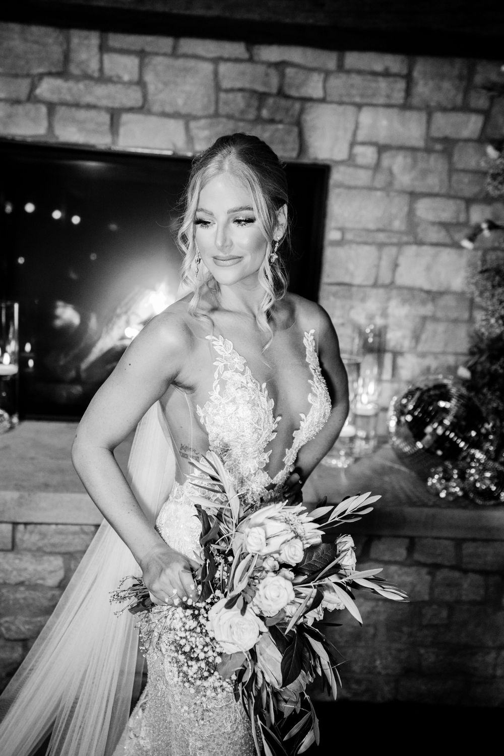 Elegant bride in lace gown holding bouquet poses by stone fireplace in black and white photo.
