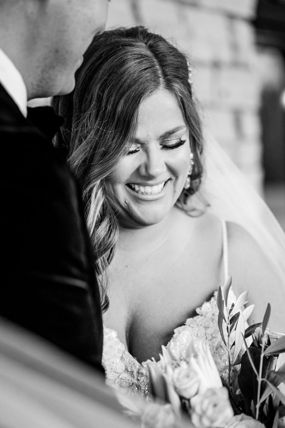 Black and white photo of a smiling bride in lace gown holding bouquet, being kissed by groom on her wedding day.