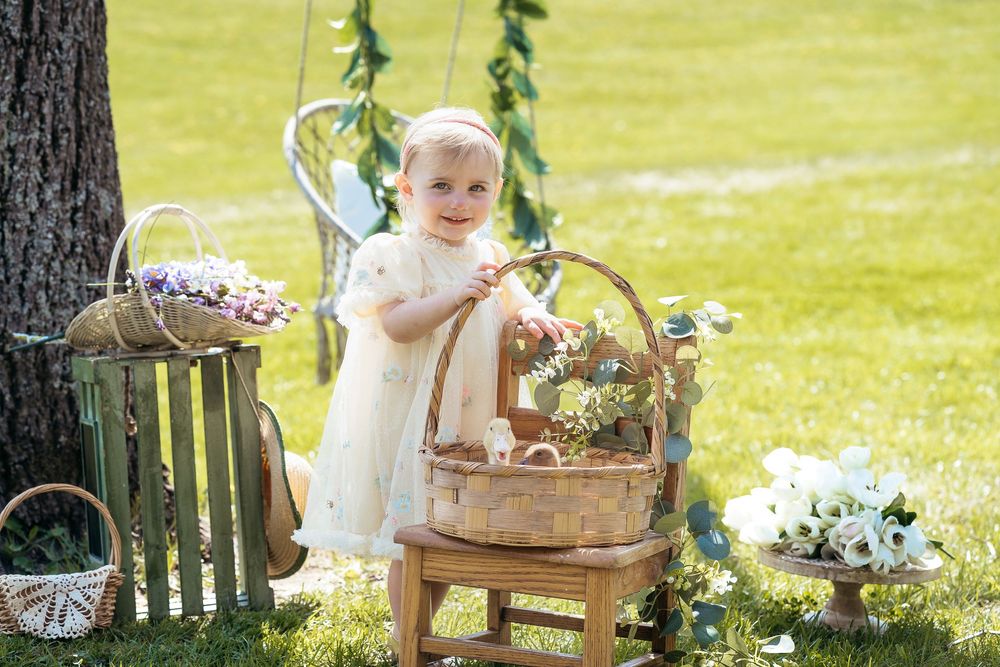 A smiling baby in a white dress stands by a wicker basket on a stool in a sunny garden with floral decorations.