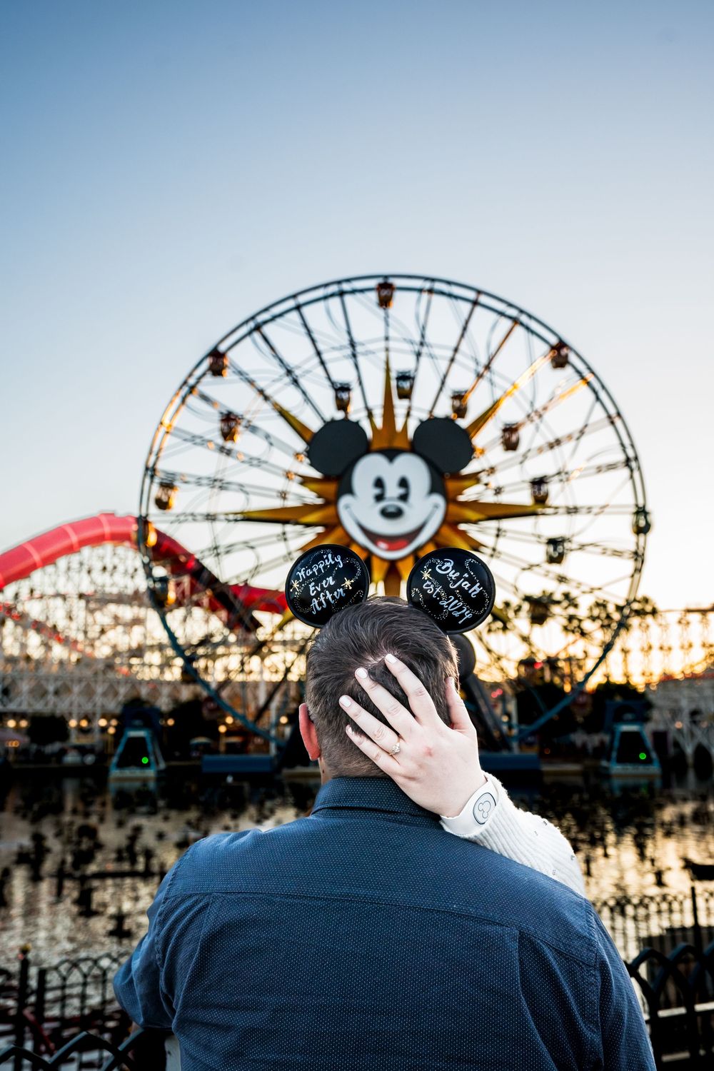 Couple embracing at Disneyland California Adventure with Mickey Mouse Ferris wheel at sunset, wearing Mickey ears.