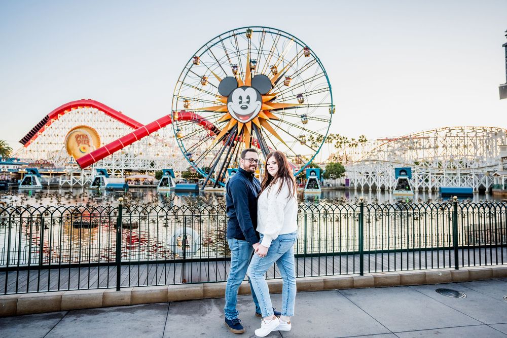Couple posing together at Disney California Adventure Park with Mickey Mouse Ferris wheel and roller coaster backdrop.