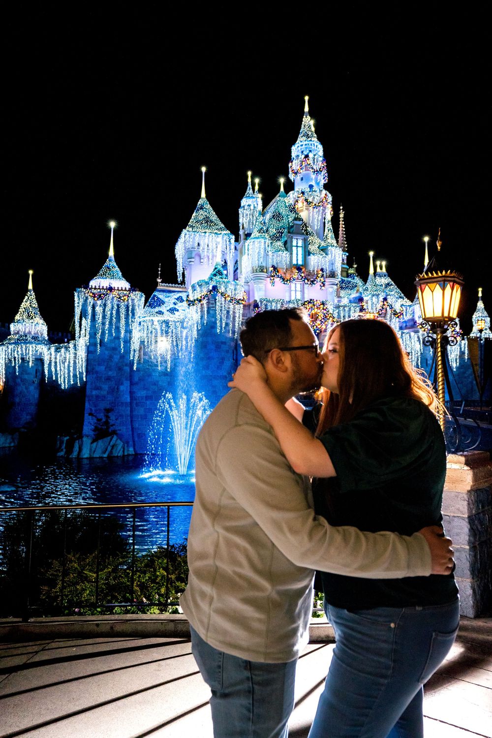 Couple sharing a romantic kiss in front of illuminated Disneyland castle at night during Christmas season.