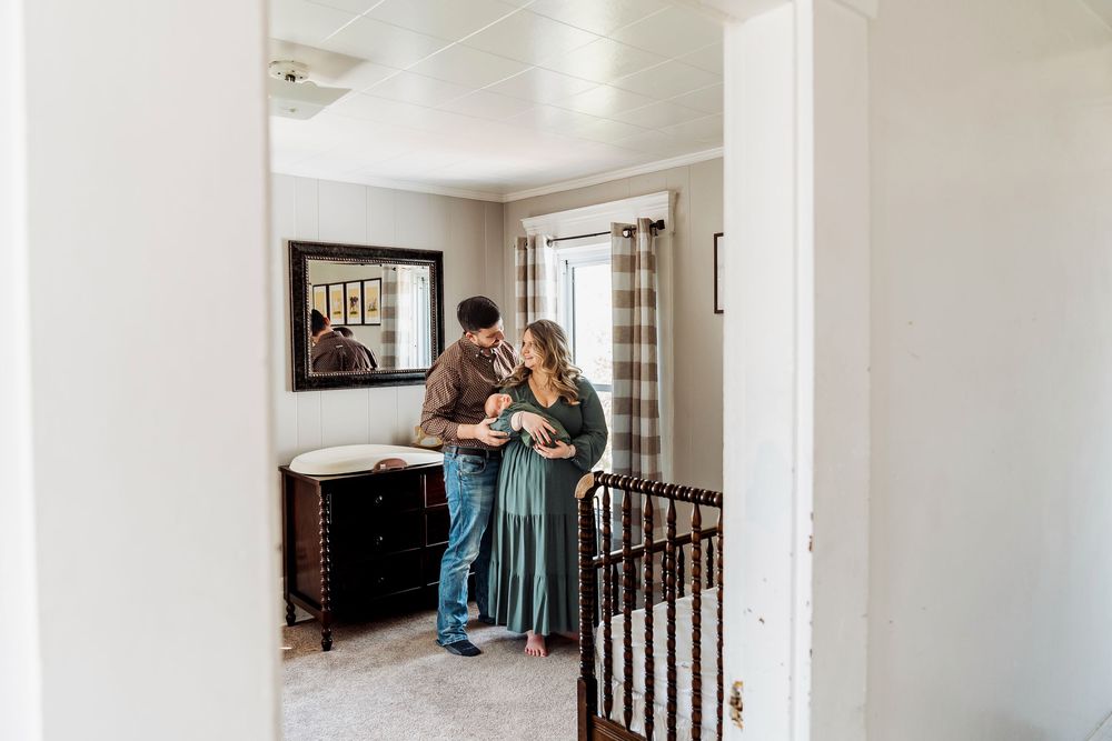 A couple stands together in a nursery, lovingly holding their newborn baby near a dresser with a mirror.
