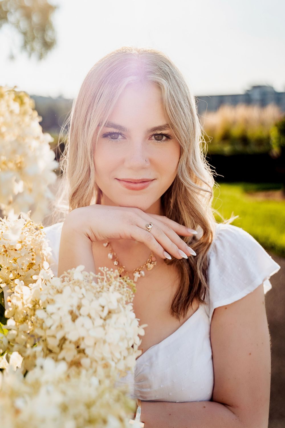 Young blonde woman in white dress holding white hydrangea flowers outdoors in golden sunlight, wearing a ring.