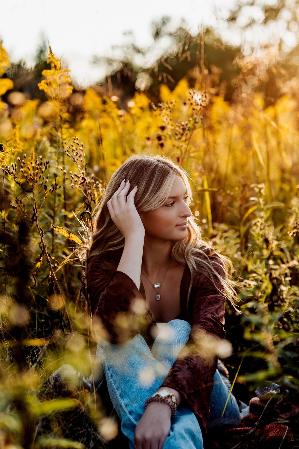 Blonde woman sitting in golden wildflower field at sunset, wearing brown jacket and jeans, hand in hair.