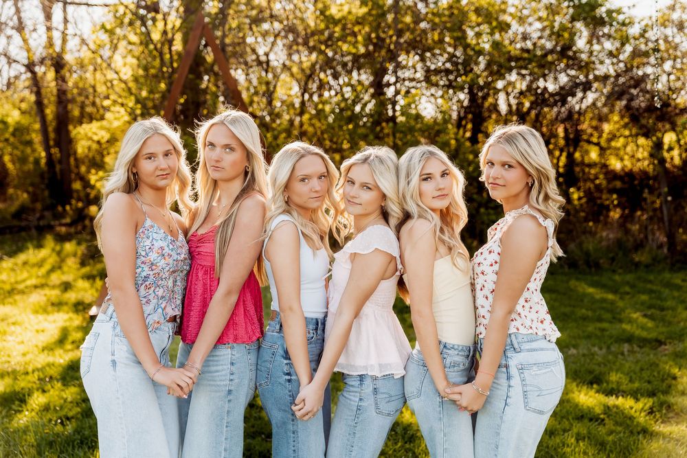 Six blonde women in jeans pose together outdoors in golden sunlight with lush green trees in the background.