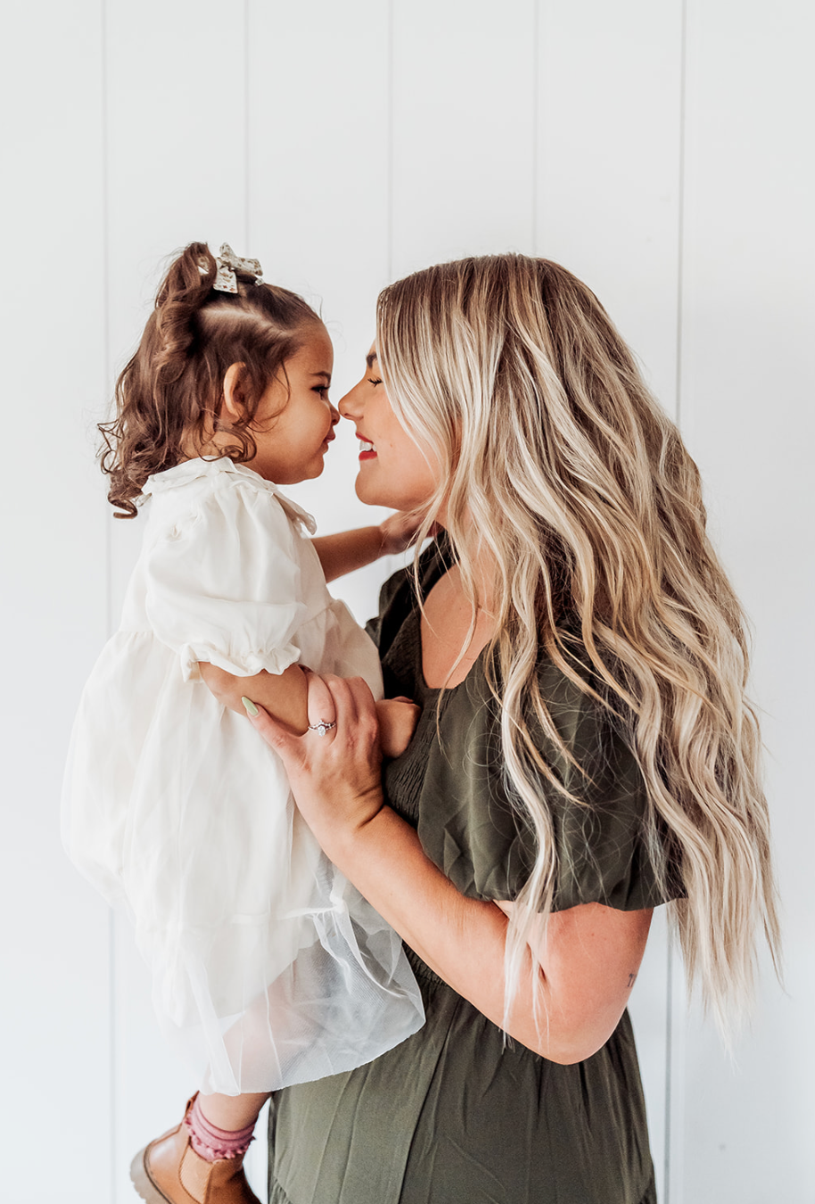 Woman with long blonde hair holding a toddler girl in a white dress, sharing a sweet nose-to-nose moment.
