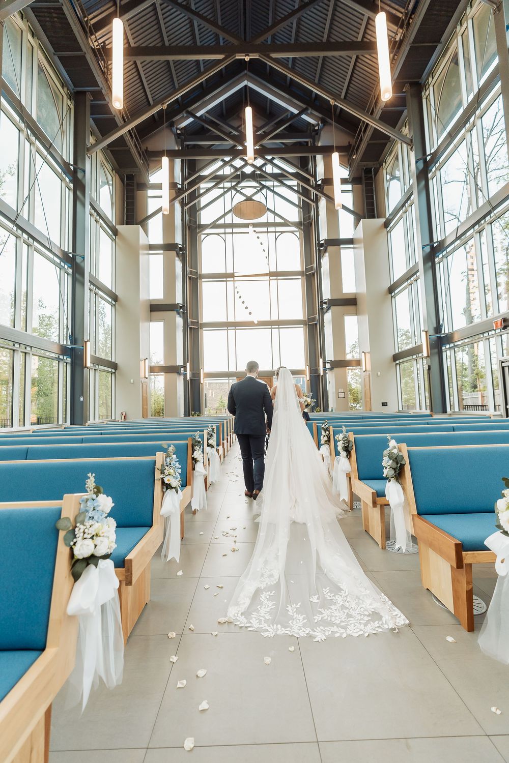 Bride and groom walk down the aisle of a modern chapel with blue pews, large windows, and a long lace wedding veil.