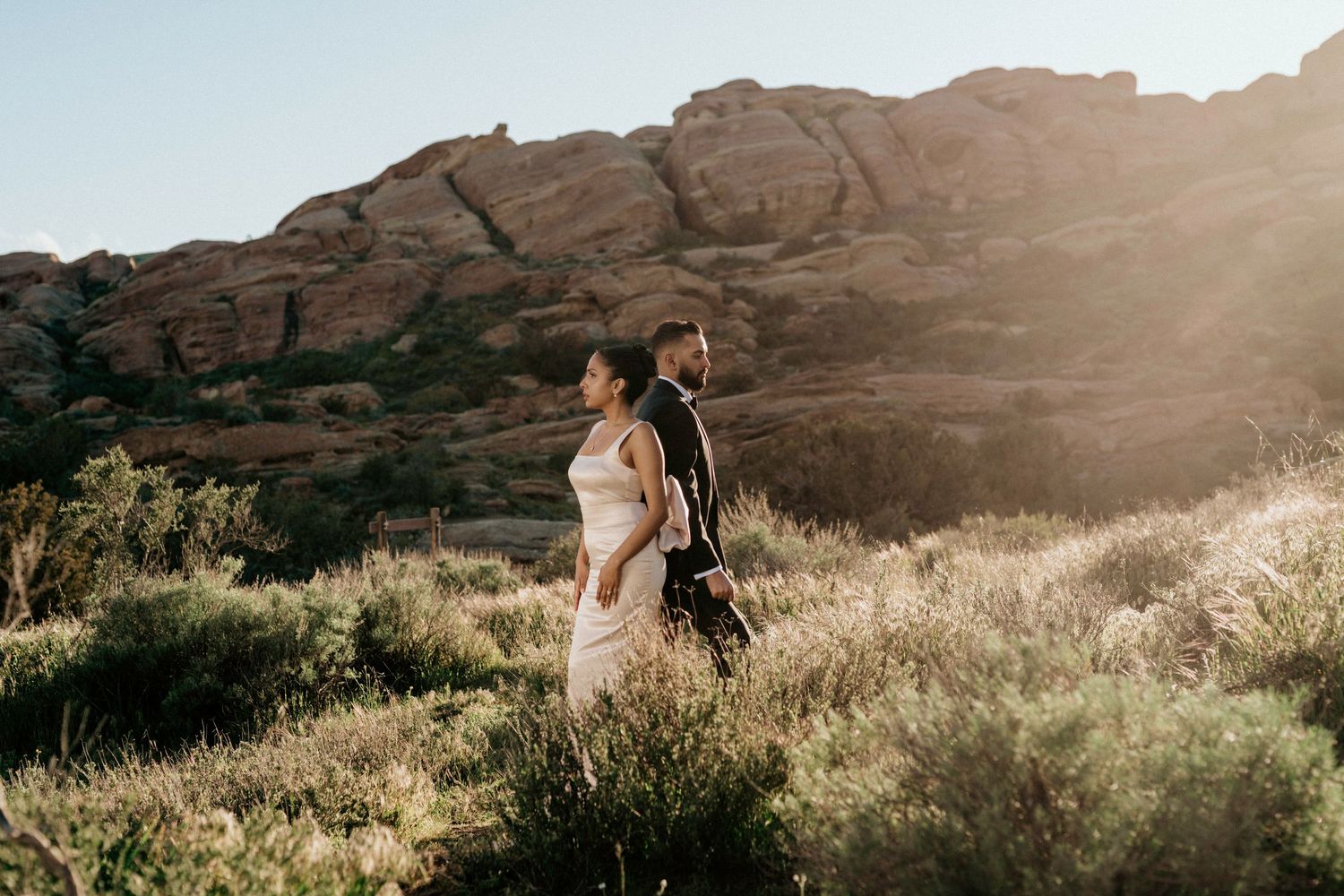A couple in wedding attire walks along dramatic rock formations at sunset in a desert landscape.