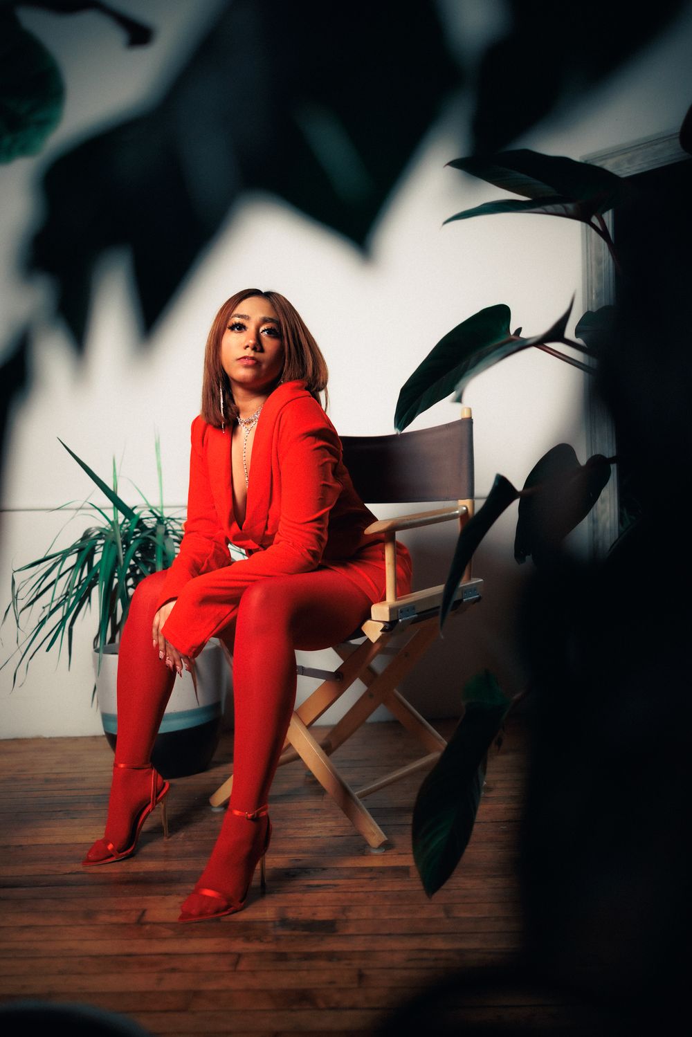 Dramatic indoor photoshoot with bright red pantsuit against plant shadows on wooden floor.