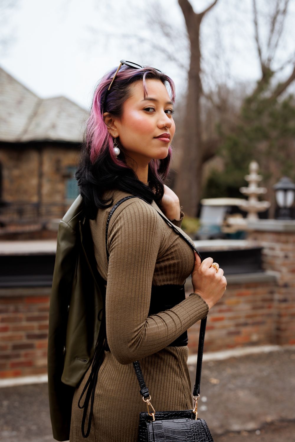A fashionable woman with pink-tinted hair wearing a brown sweater and vest poses outdoors near a brick wall.