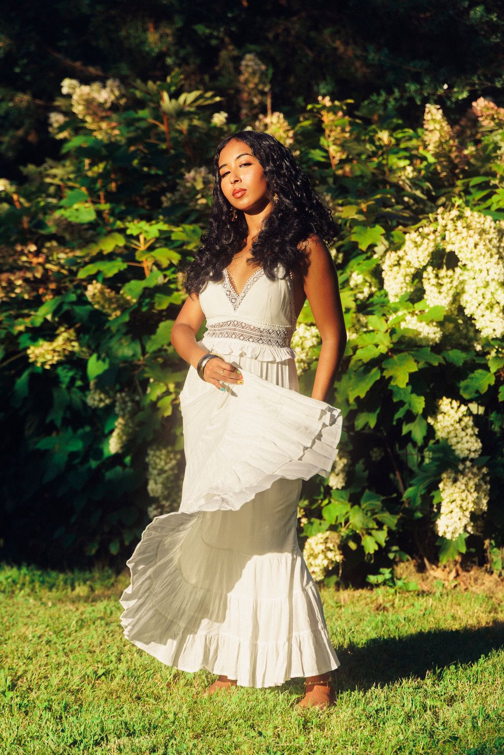 A person in a flowing white dress poses gracefully in a garden setting with blooming hydrangea bushes in the background.