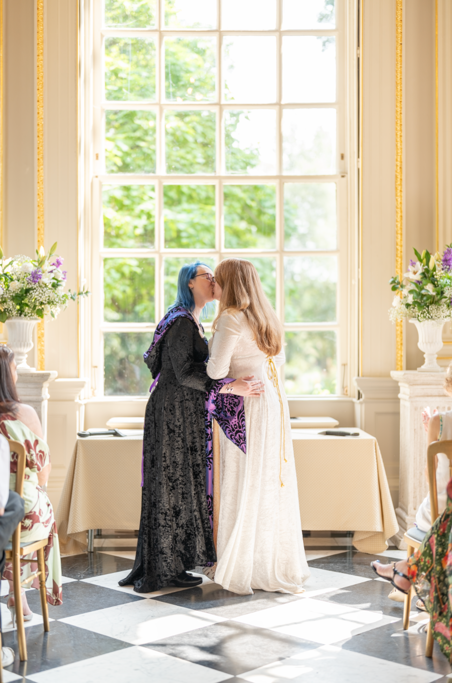 Wedding couple sharing kiss by sunlit window in elegant room with floral arrangements.