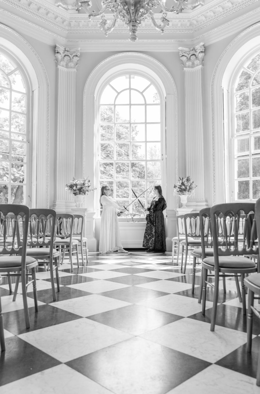 Elegant white room with arched windows, checkered floor and rows of chairs set up for a ceremony.