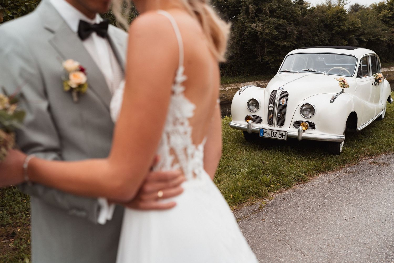 Brautpaar steht eng beisammen vor einem weißen Oldtimer, während die Braut ihren Blumenstrauß hält