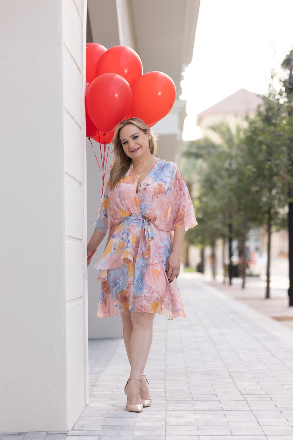 A person in a floral summer dress poses with red heart balloons on a sidewalk near white columns.