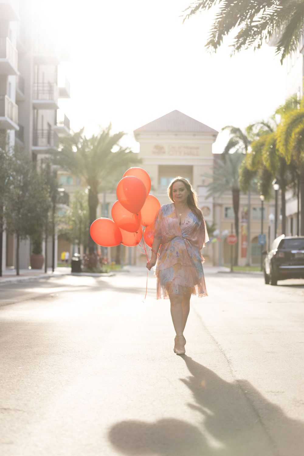 A person in a flowy dress skips down a palm tree-lined street holding orange balloons on a sunny afternoon.