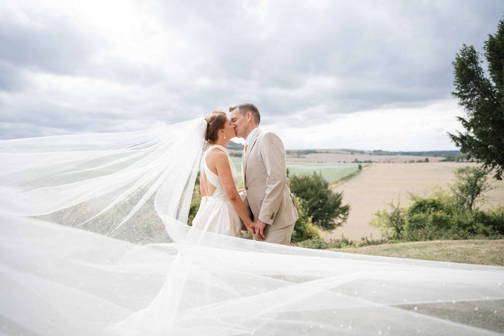 A couple shares a romantic kiss on a sandy beach with a flowing white veil caught in the breeze.