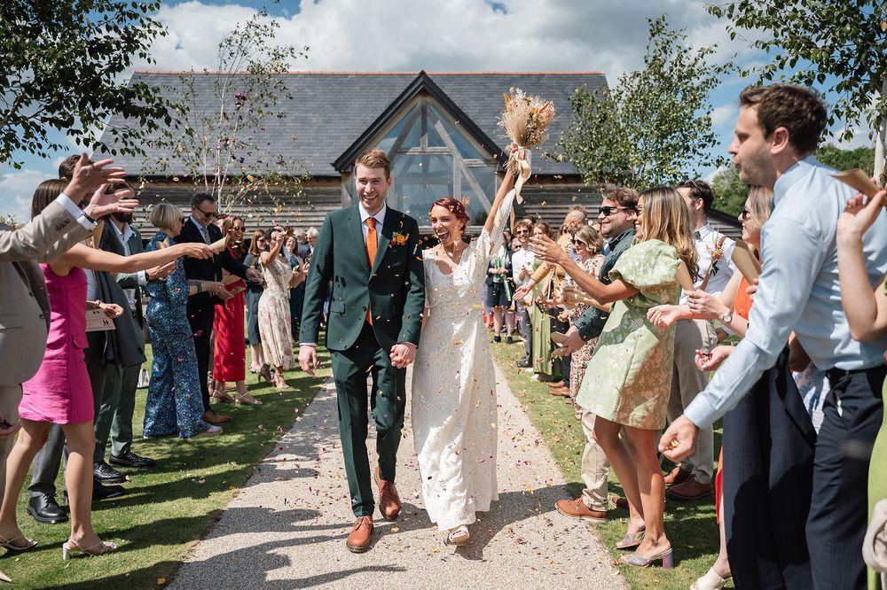 Newlyweds walking through crowd of celebrating guests throwing confetti outside rustic venue.