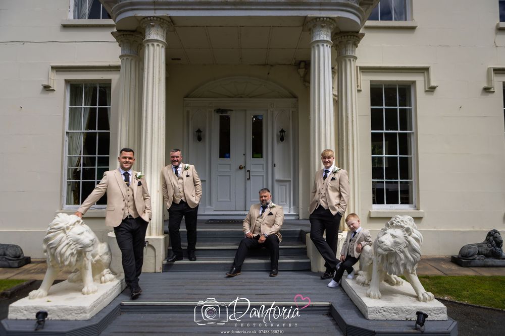 Groom and groomsmen in champagne suits pose on steps of elegant manor house with stone lion statues.