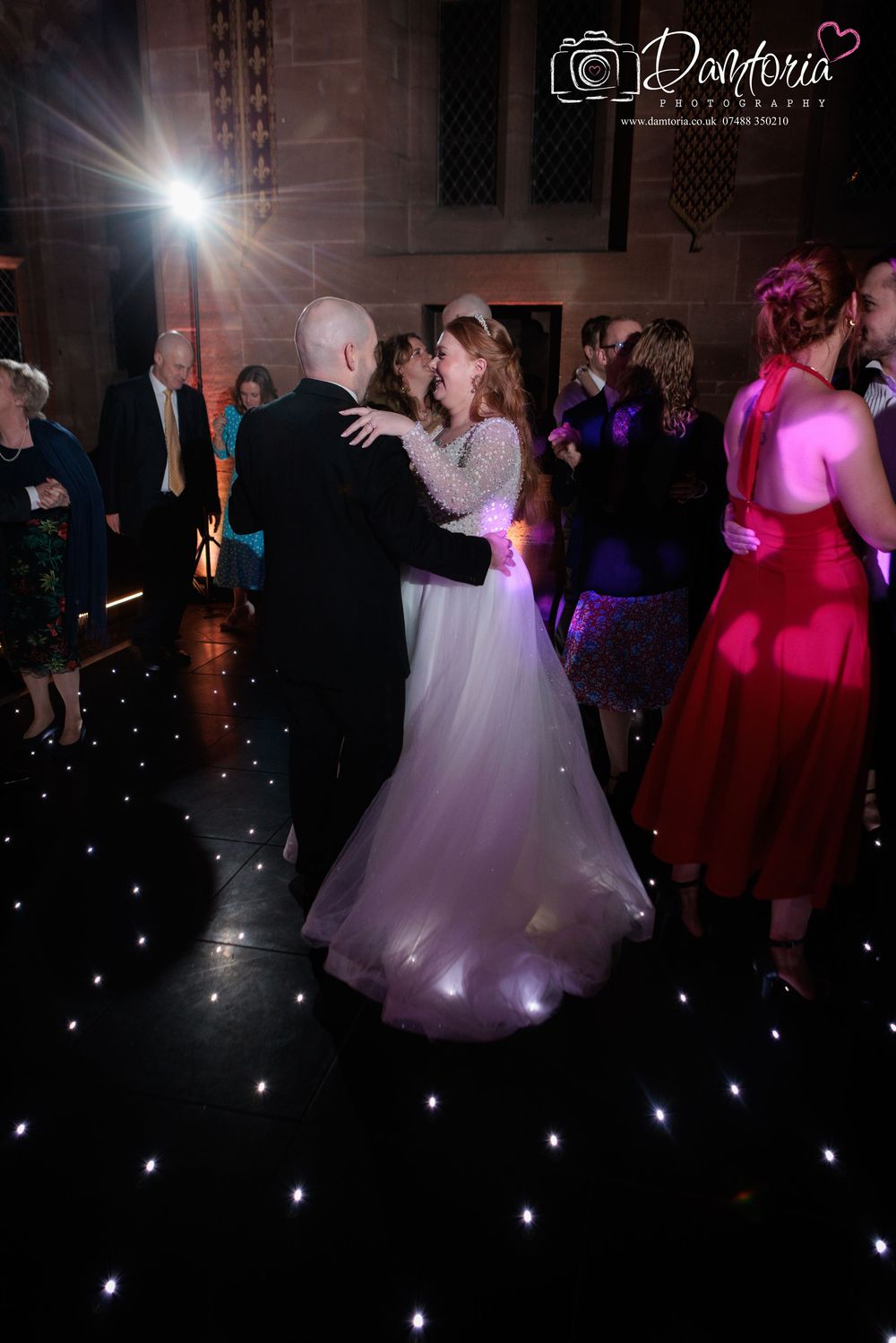 Wedding guests dance on a dark dance floor illuminated by twinkling lights and colorful spotlights during a reception celebration.