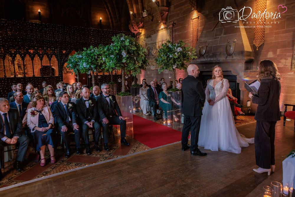A wedding ceremony takes place in a grand historic hall with stone arches and dramatic lighting.