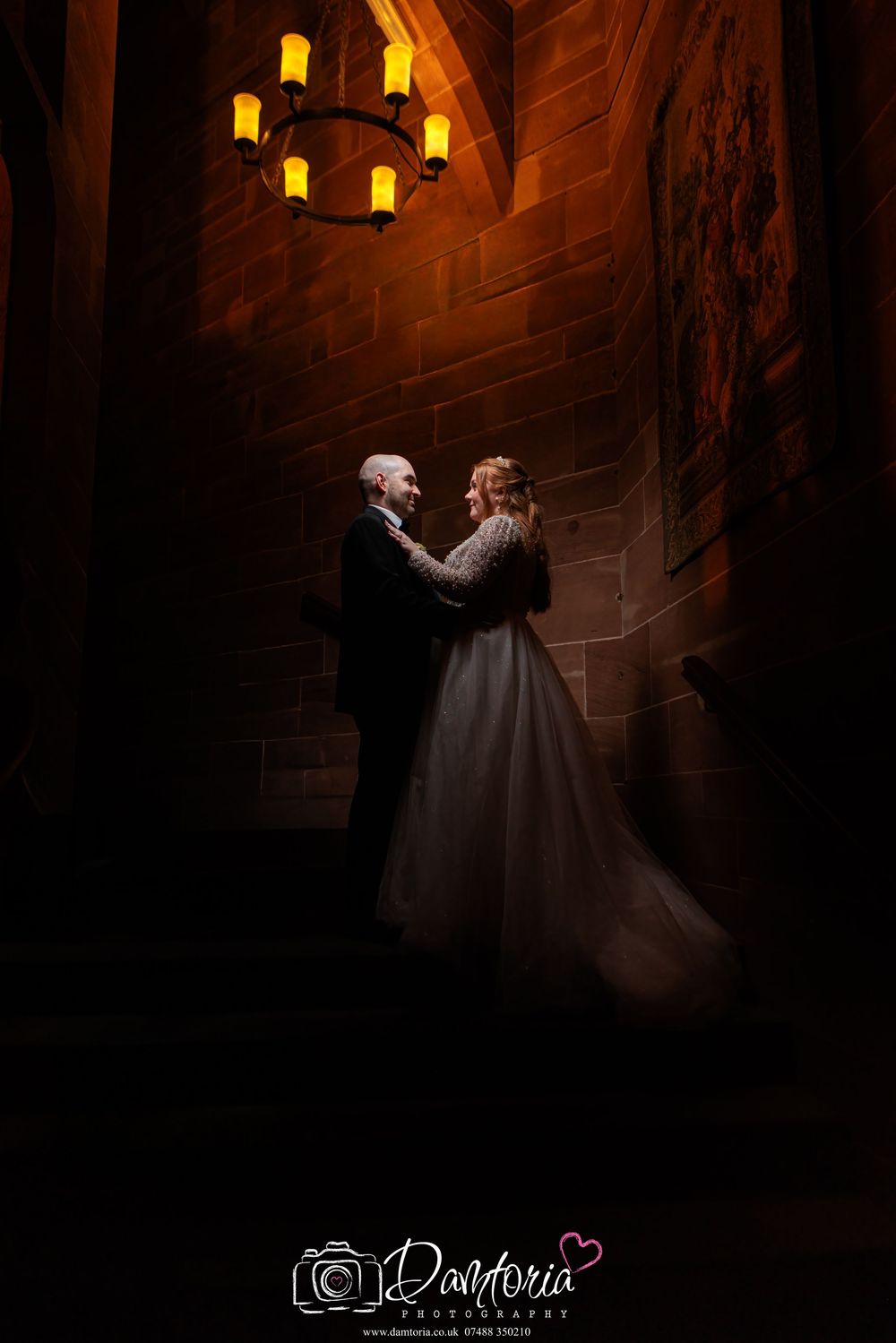 A romantic couple embraces in a dimly lit stone hallway with a glowing chandelier overhead casting warm light.