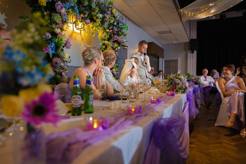 Wedding reception head table with floral arch, purple decorations, candles, and groom giving a speech to seated guests.