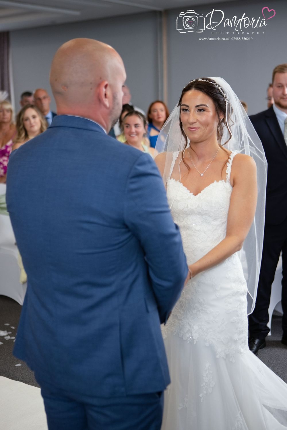 Bride in lace wedding dress and veil smiles at groom in blue suit during wedding ceremony with guests seated behind.