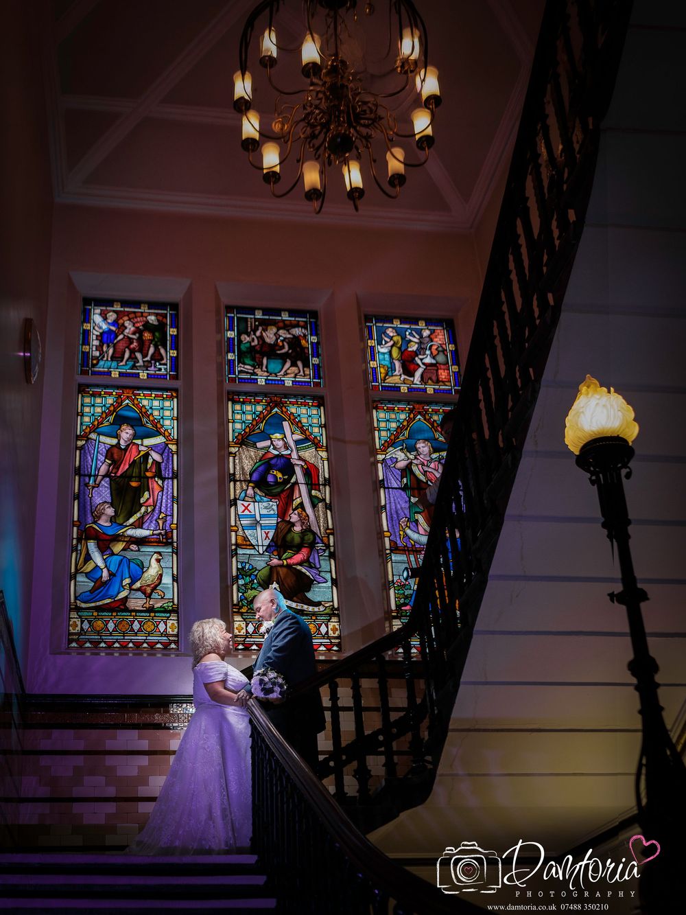 Bride and groom on grand staircase with stunning stained glass windows and chandelier at elegant wedding venue.