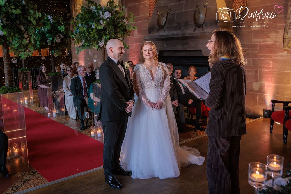 Wedding ceremony guests gather inside a rustic wooden barn venue illuminated by warm lighting and decorated with greenery.