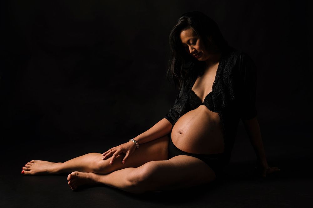 Artistic maternity photo with dark backdrop shows silhouette in black attire sitting gracefully on floor.