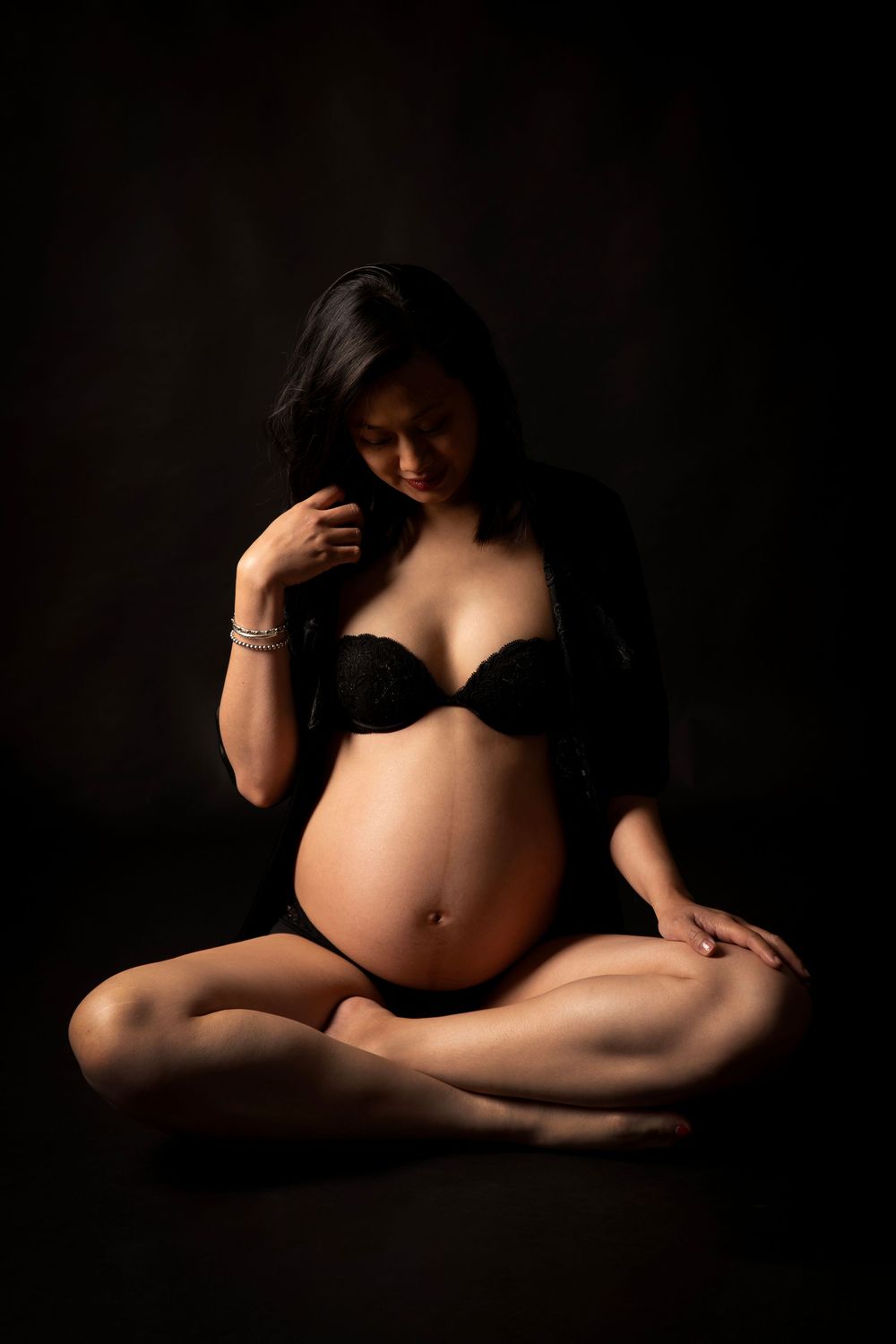 Artistic maternity photo showing an expectant mother in black lingerie sitting cross-legged against dark background.