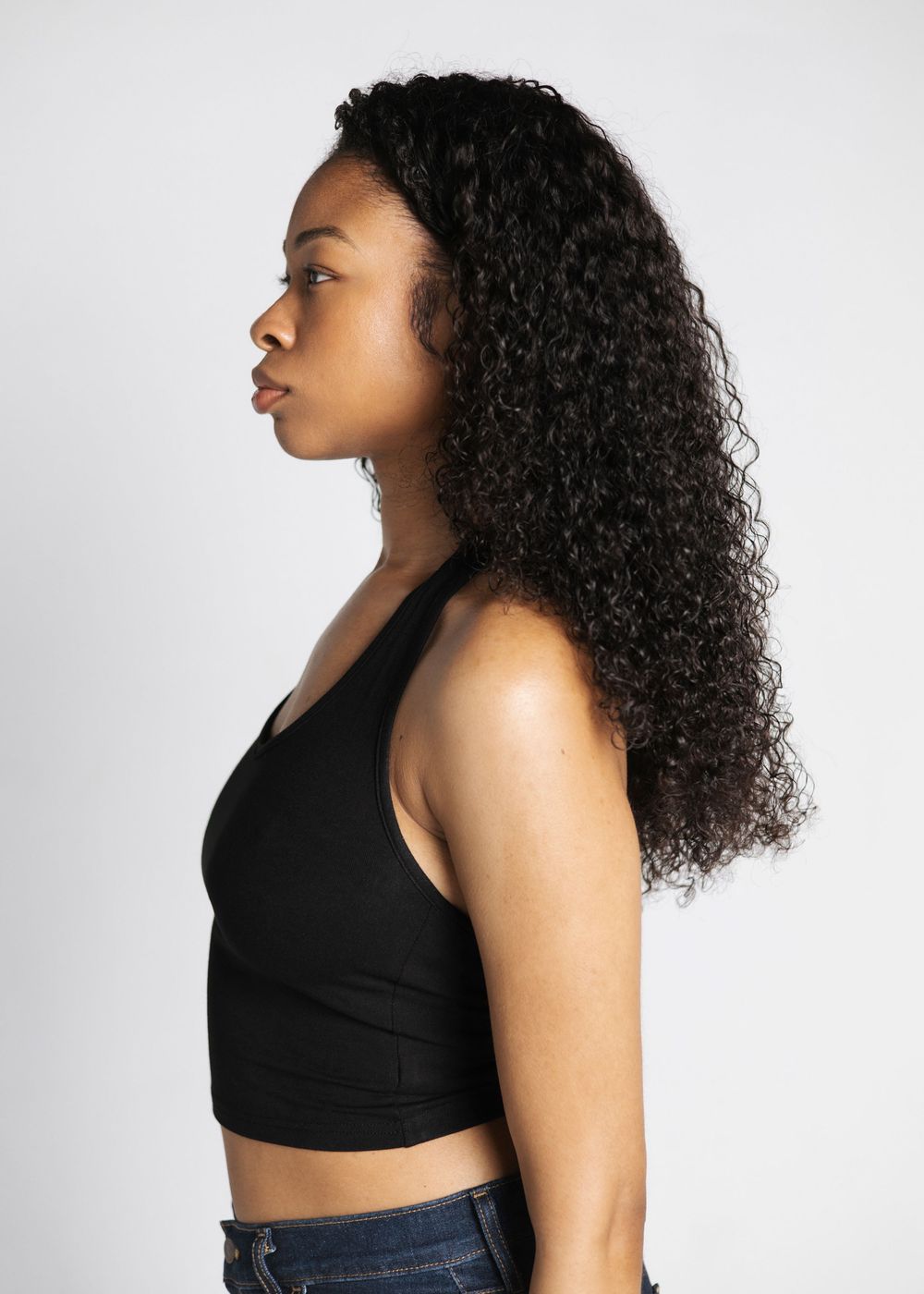 Woman with long curly hair wearing a black crop top and jeans, photographed from the side against a white background.