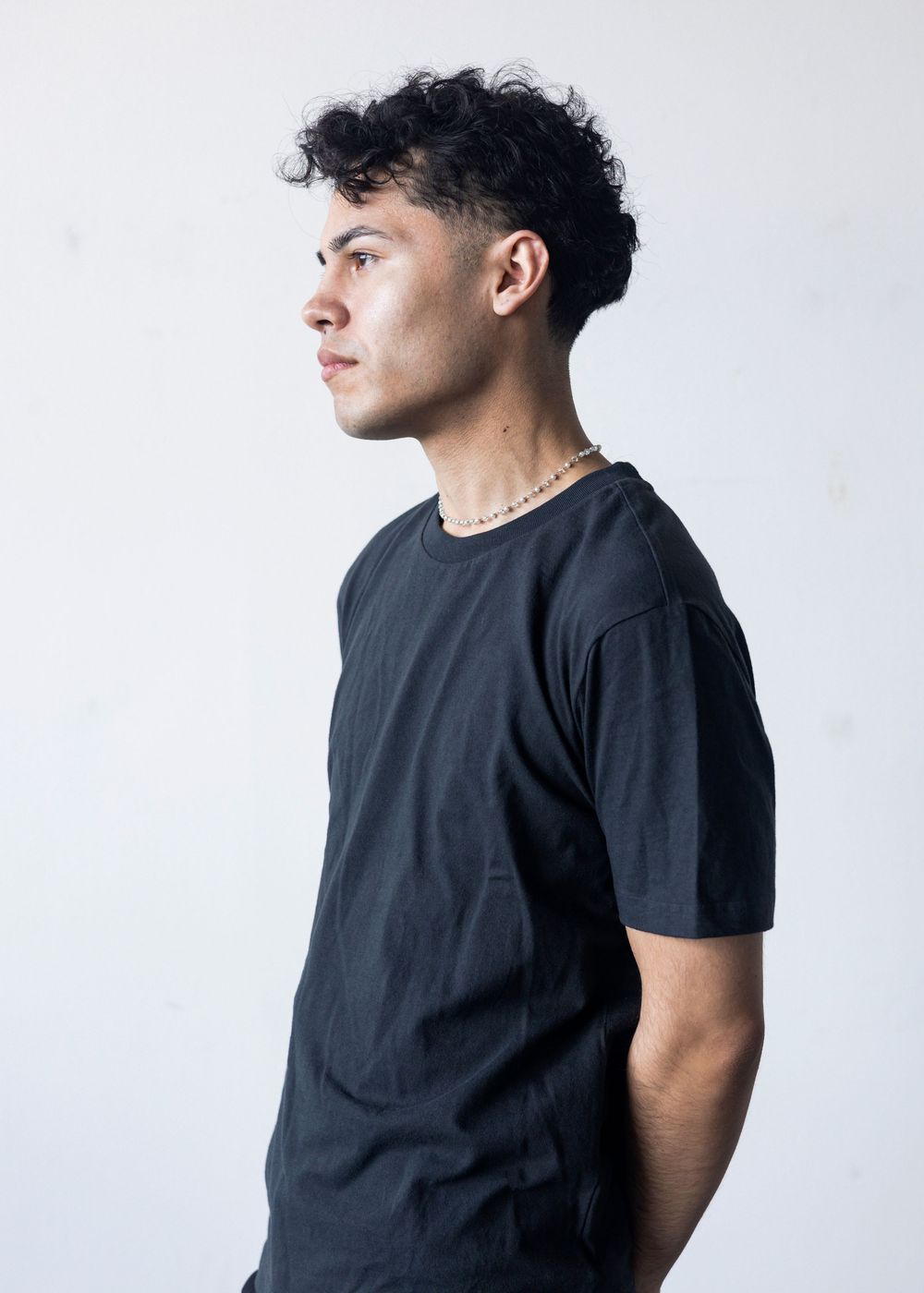 Young man in black t-shirt with curly hair and gold chain necklace posing in profile against white background.