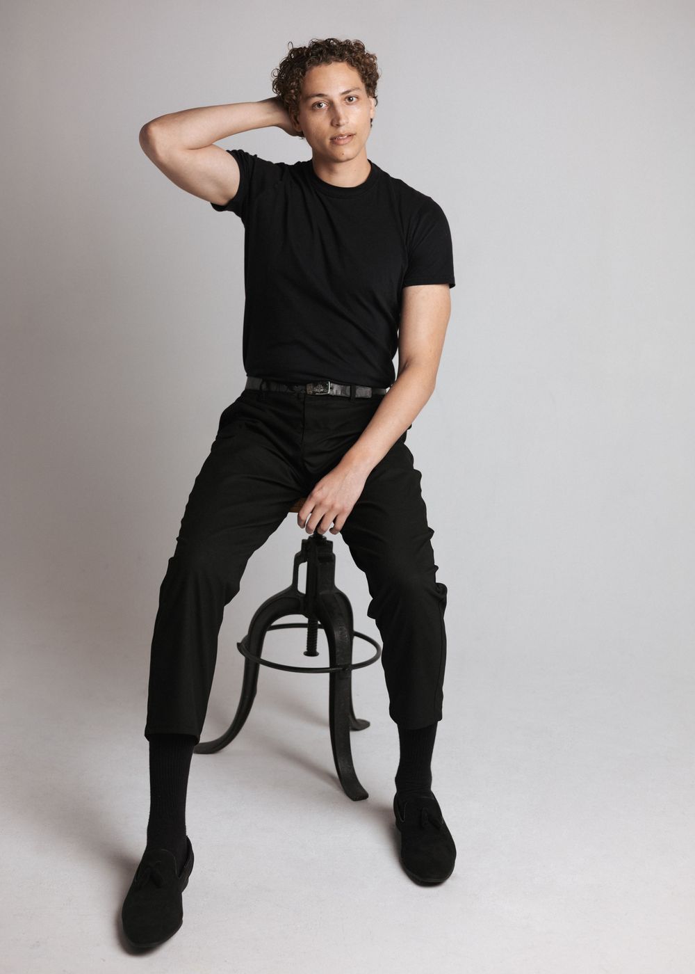 Person in all black outfit strikes casual pose while sitting on dark stool against light grey backdrop.
