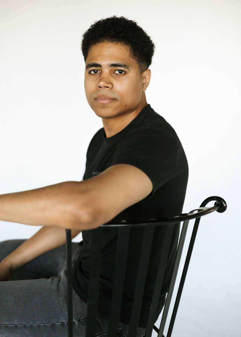 Young man in black t-shirt sits sideways on metal chair, looking over shoulder against white background.