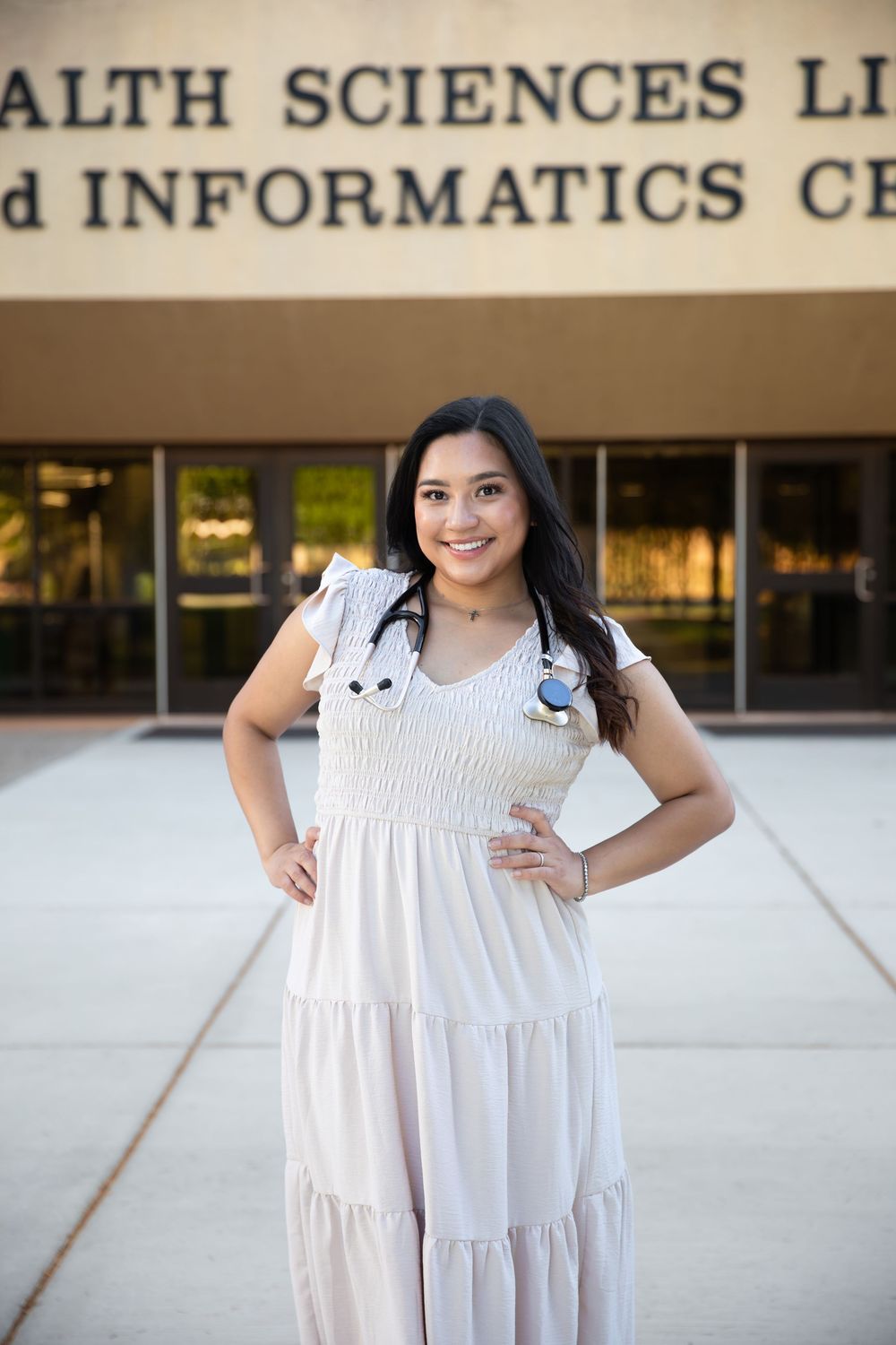 Young woman in white dress with stethoscope stands confidently outside Health Sciences Library and Informatics Center.