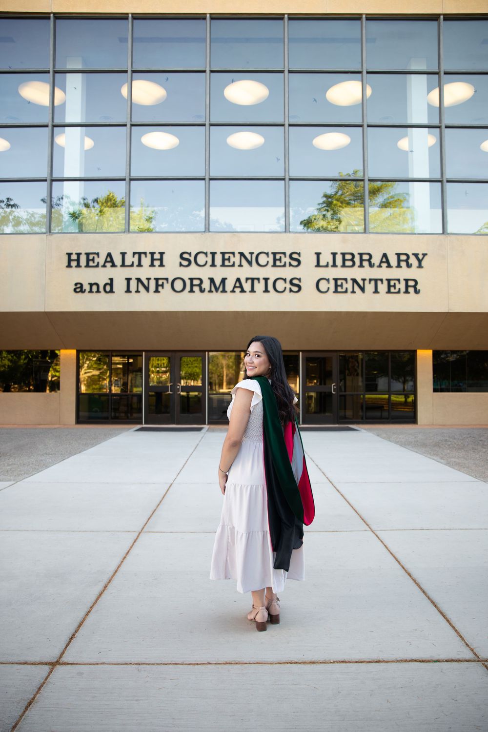 Graduate in white dress holding academic regalia outside Health Sciences Library and Informatics Center.