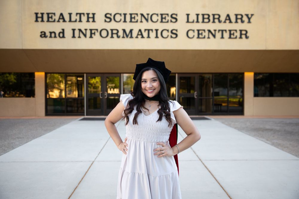 Graduate in cap and gown poses proudly outside Health Sciences Library and Informatics Center building.